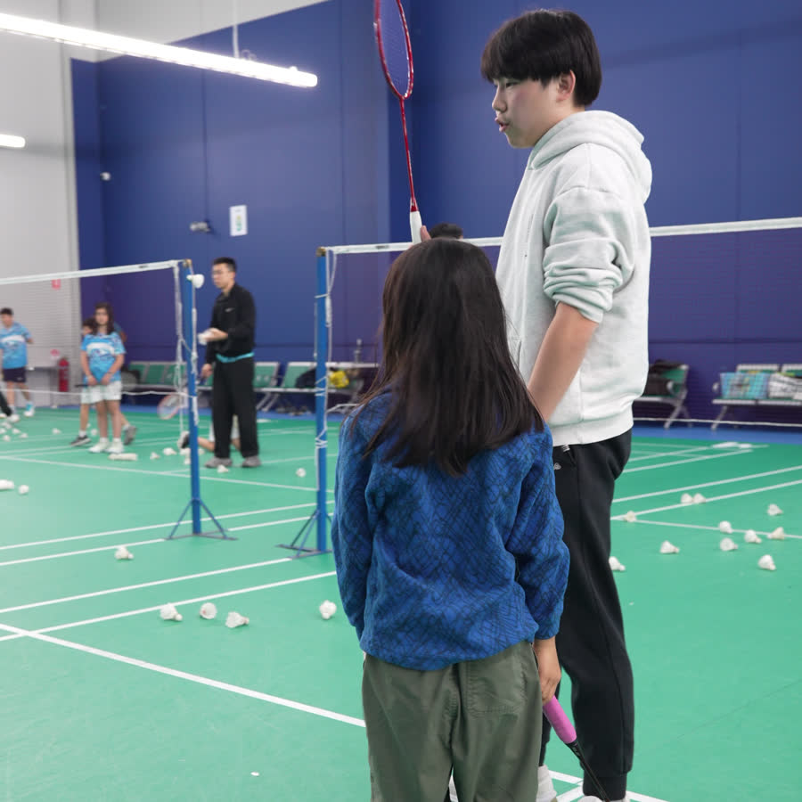 A Go Badminton coach on court beside a young girl holding a racket, showing her how to set up for a shot, with shuttles scattered across the floor.