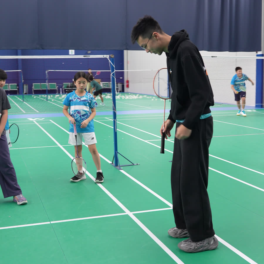A small group of children on an indoor badminton court with a coach, each with a racquet.