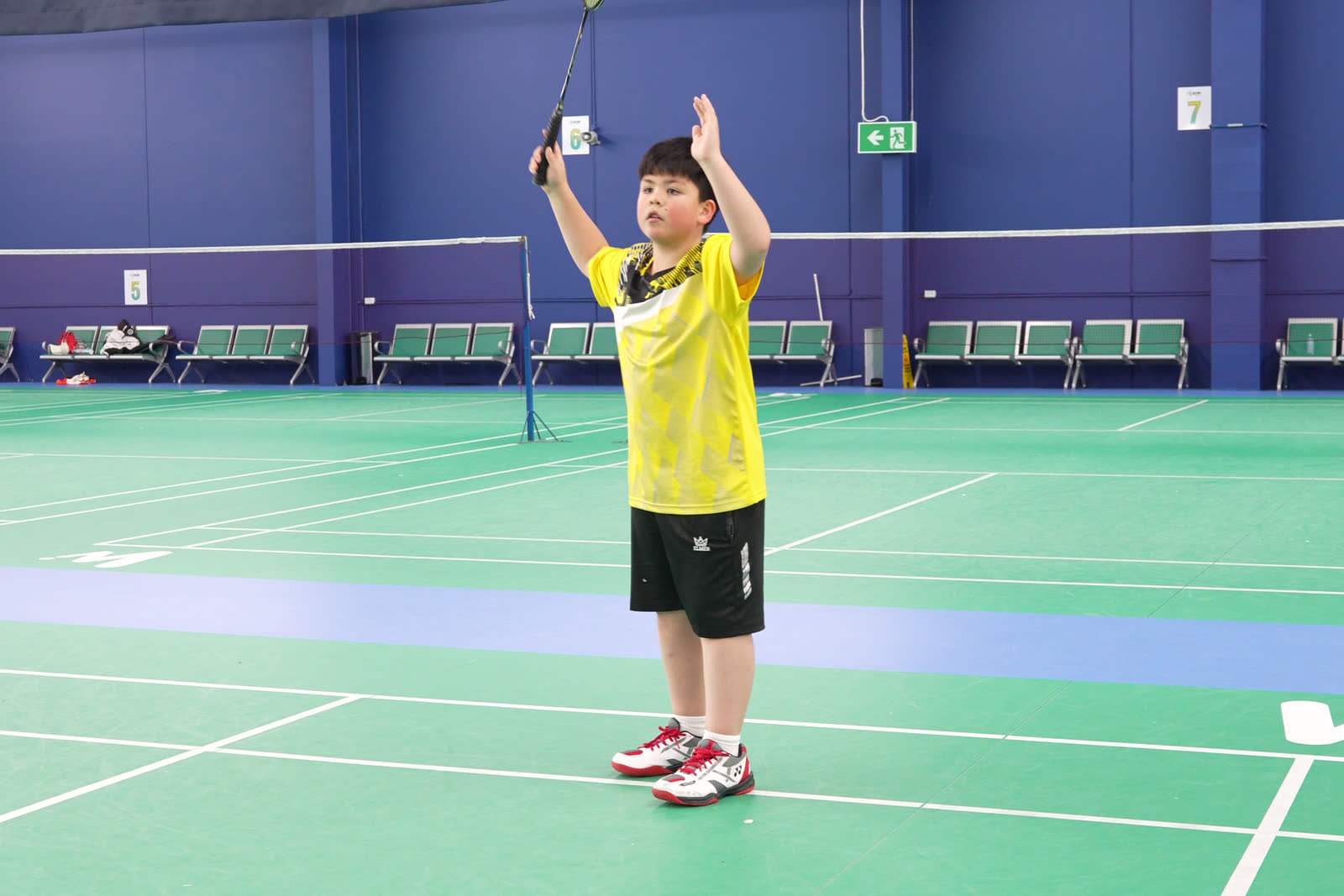 A boy in a yellow shirt reaching up to hit an overhead shot on an indoor court.